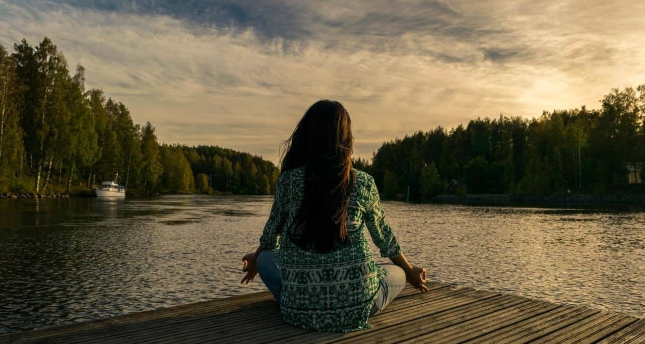 Mulher sentada em um deque, de costas, olhando o lago ao entardecer